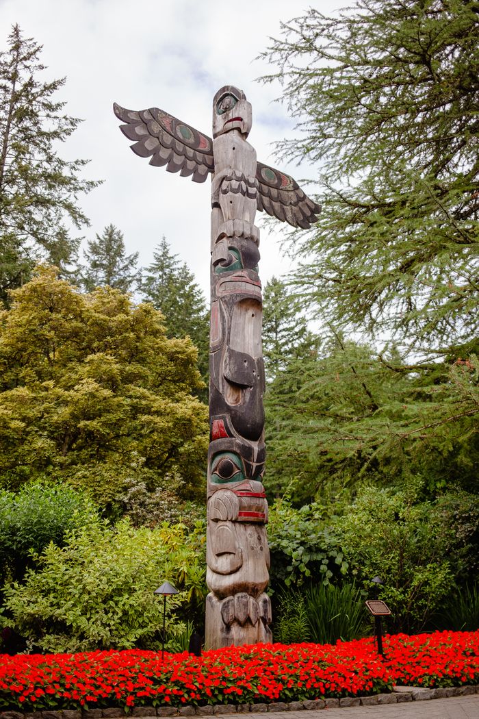A Coast Salish totem pole stands amid vibrant red flowers and lush green trees, captured from a low angle.