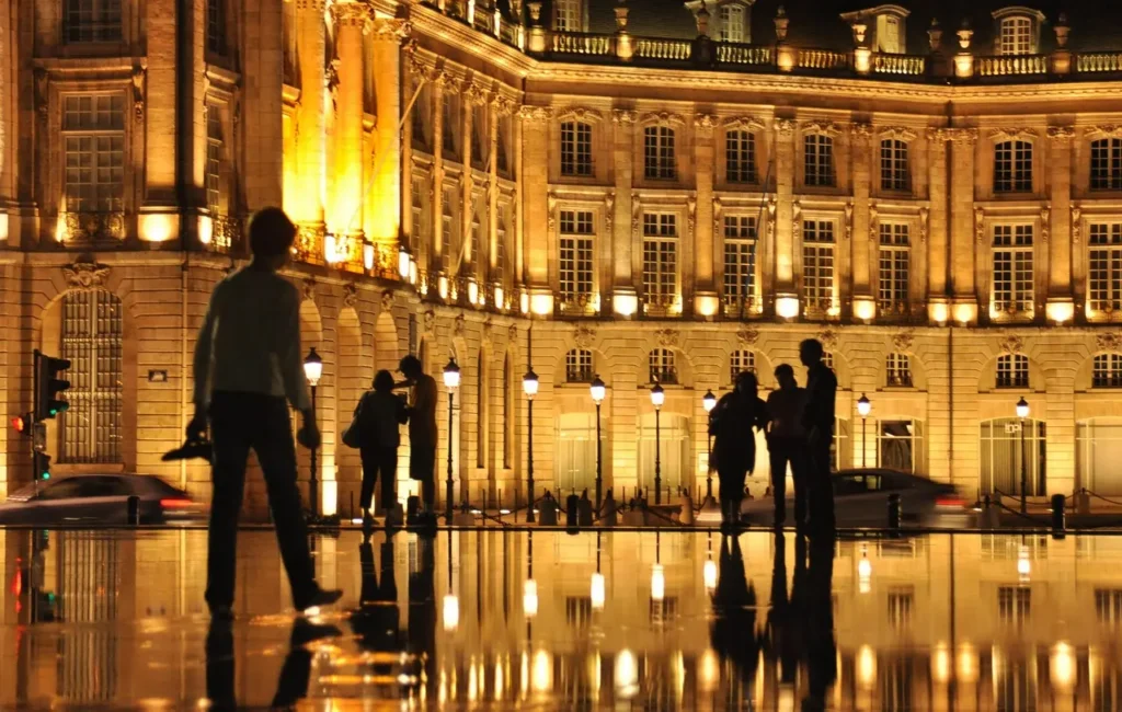 People walking beside the illuminated Place de la Bourse reflected in the Miroir d'Eau at night in Bordeaux