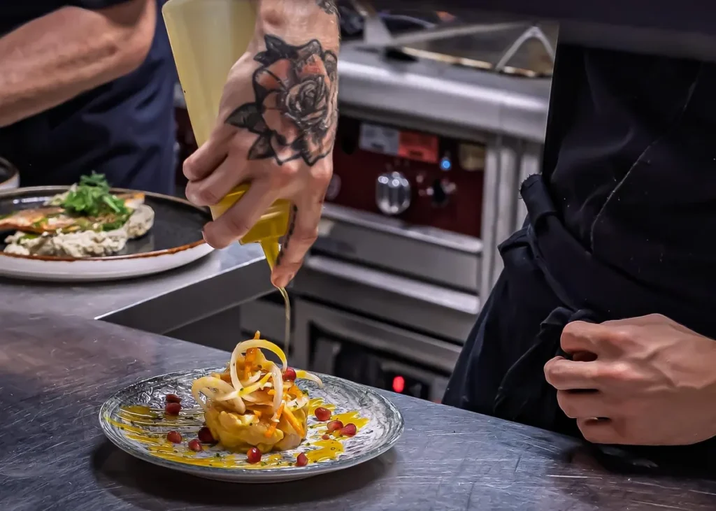 Chef adding olive oil to a freshly prepared Mediterranean dish in a restaurant kitchen