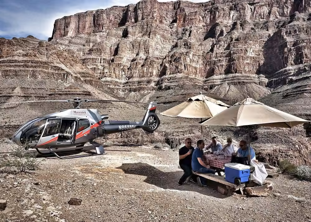 Maverick helicopter landed inside the Grand Canyon with travelers enjoying refreshments beneath towering canyon cliffs