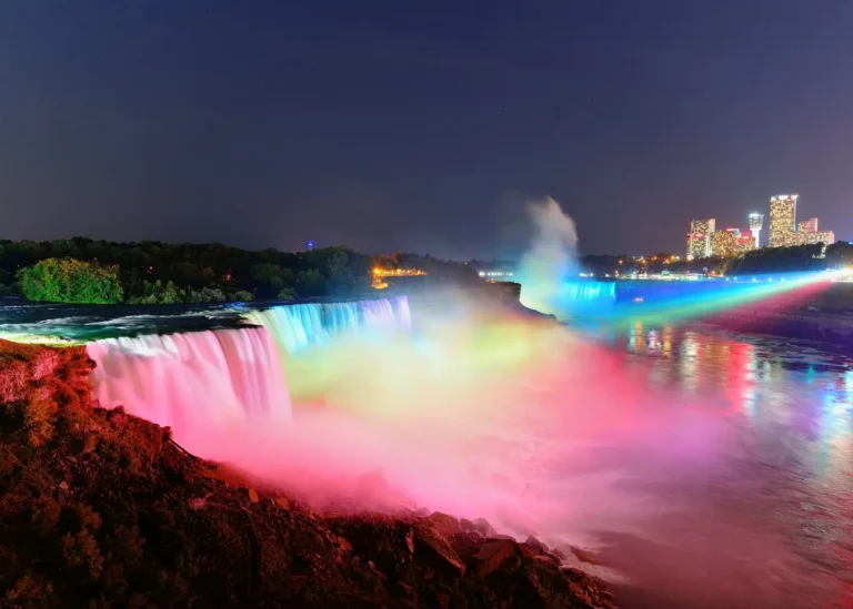 Niagara Falls illuminated with colorful rainbow lights at night, with American Falls in the foreground and Horseshoe Falls in the distance