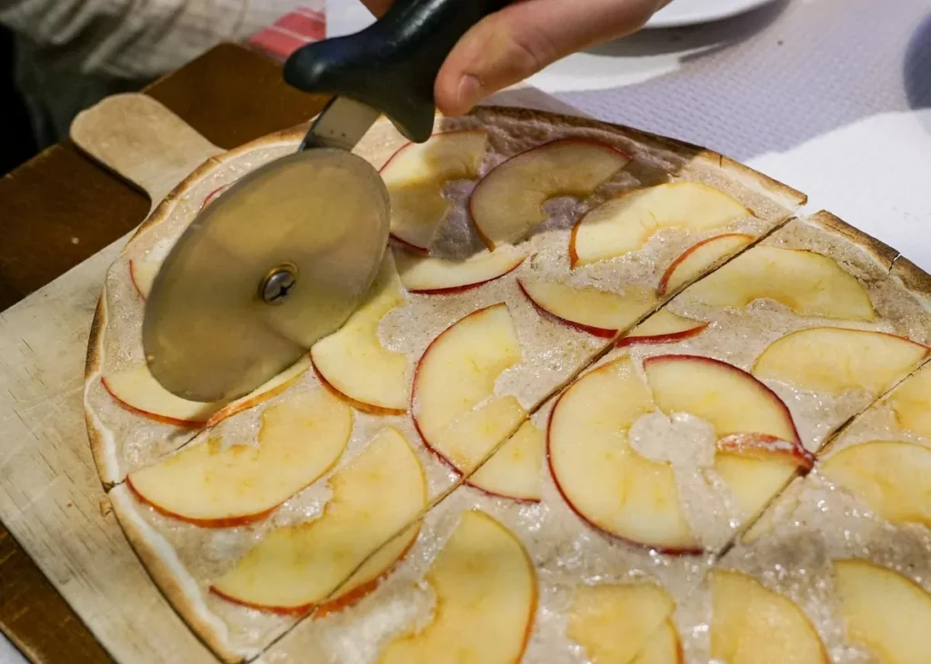 Apple tarte flambée topped with thin apple slices being cut with a pizza wheel on a wooden board