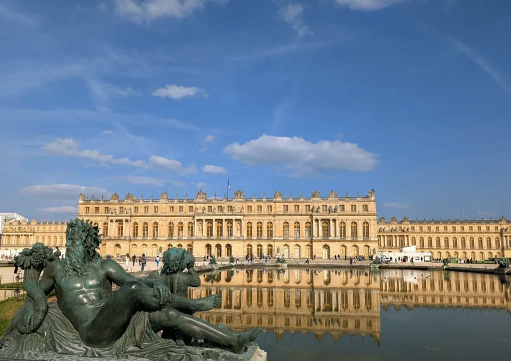 Palace of Versailles garden facade reflected in a fountain, with a bronze sculpture in the foreground